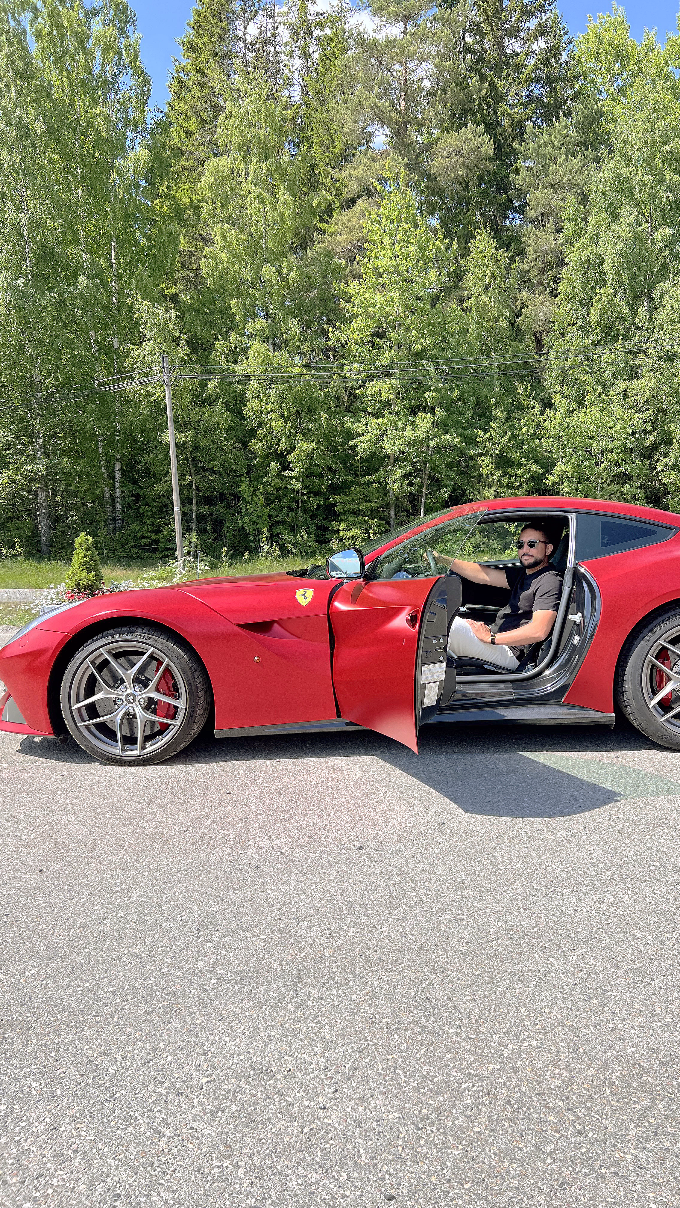 Mann i solbriller sitter i en rød Ferrari med åpen dør foran en skog