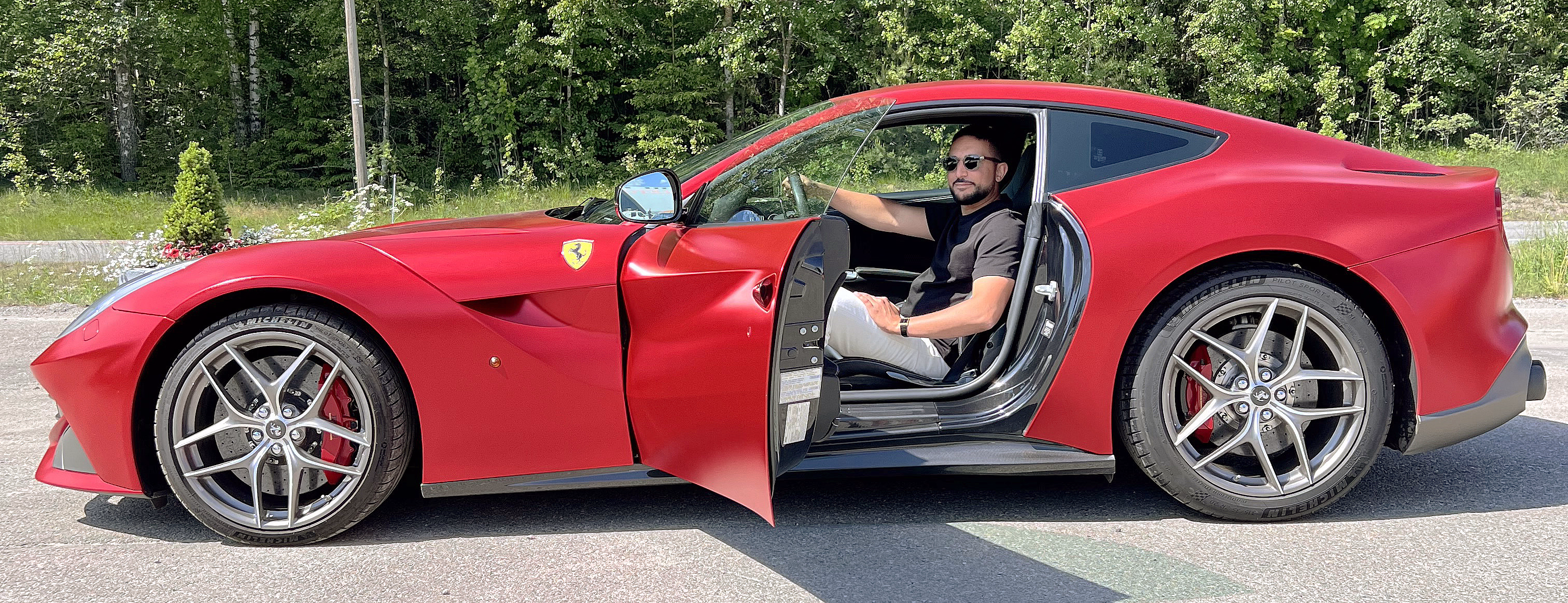 Mann i solbriller sitter i en rød Ferrari med åpen dør foran en skog