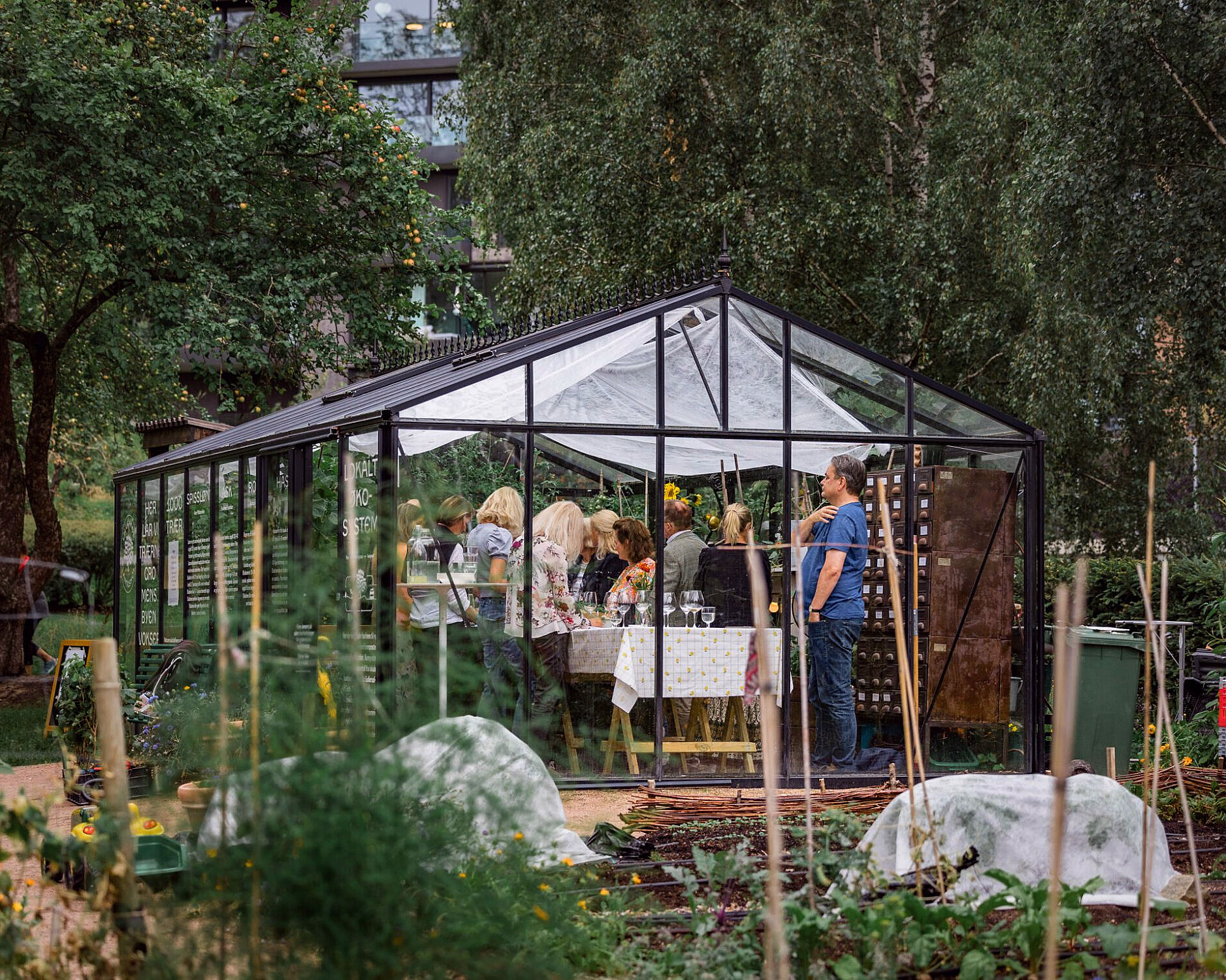 Folk samlet rundt et bord inne i et drivhus av glass og svart metall i en hage