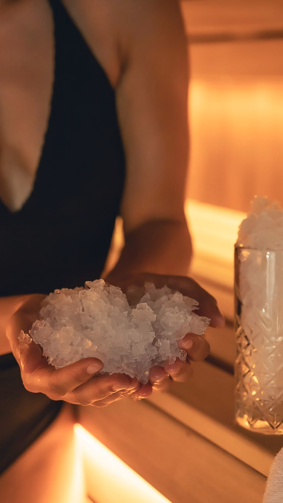 Person i sort badedrakt holder knust is i hendene ved siden av et glass med is og et hvitt håndkle
