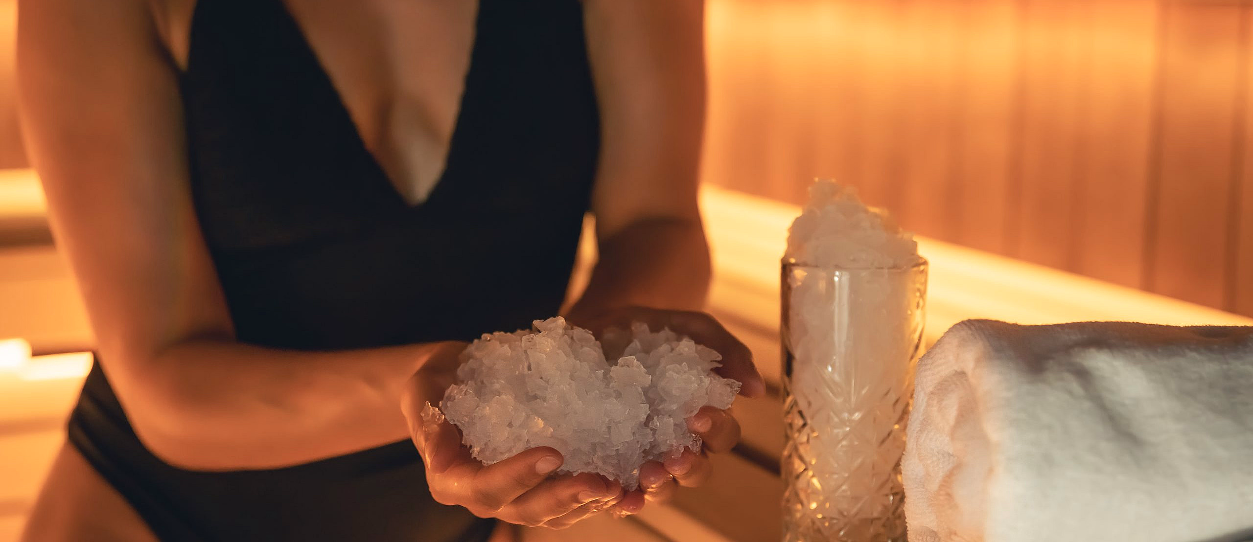 Person i sort badedrakt holder knust is i hendene ved siden av et glass med is og et hvitt håndkle