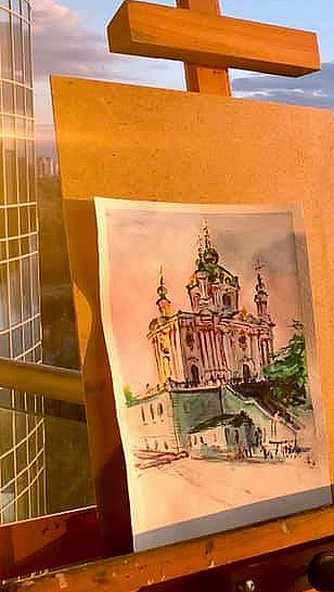 Maleri av en fargerik katedral på et staffeli på en balkong ved en elv i solnedgang