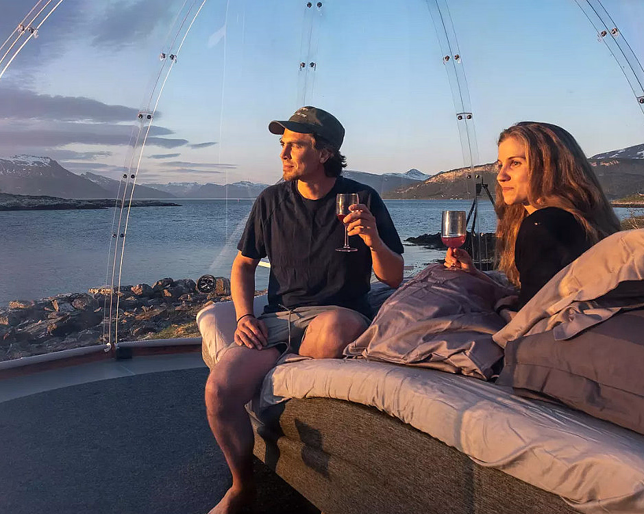 Mann og kvinne i en gjennomsiktig kuppel med glass i hendene, utsikt over vann og fjell