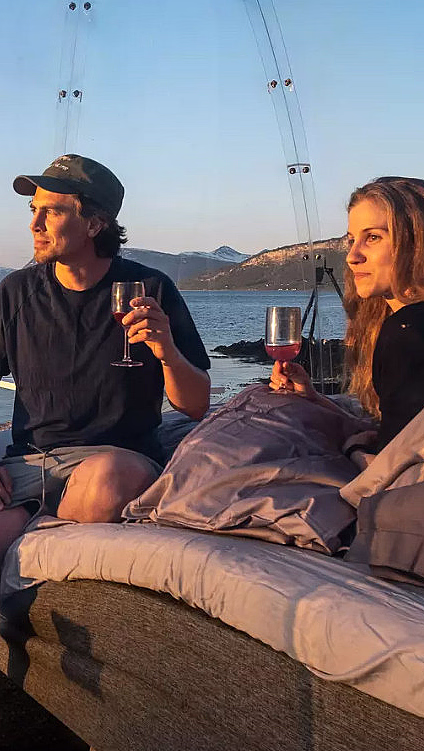 Mann og kvinne i en gjennomsiktig kuppel med glass i hendene, utsikt over vann og fjell