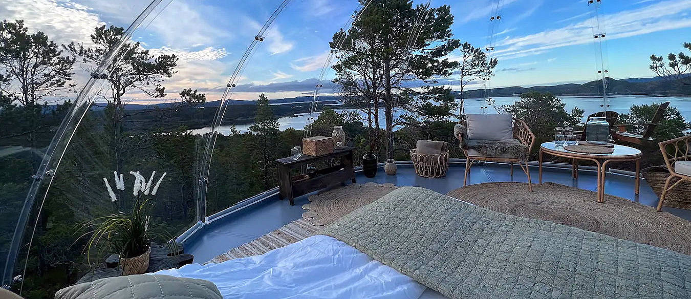 Gjennomsiktig kuppel med seng og stoler med utsikt over vann og skog