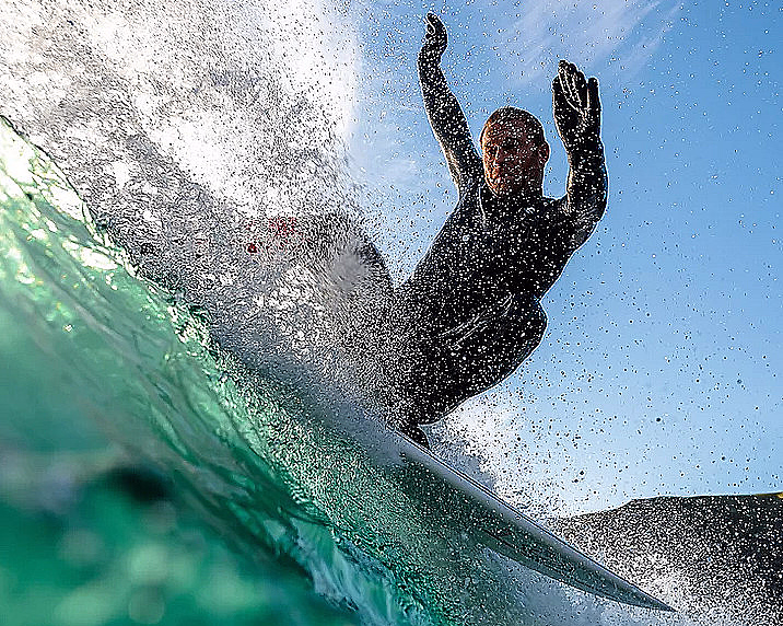 Surfer i svart våtdrakt rir på en bølge med armene hevet mot en blå himmel