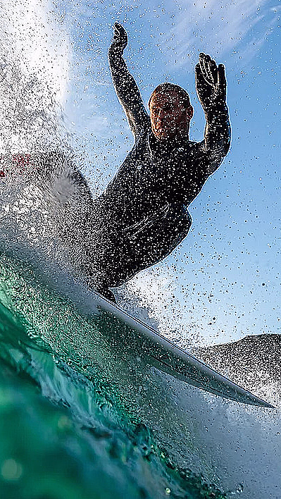 Surfer i svart våtdrakt rir på en bølge med armene hevet mot en blå himmel