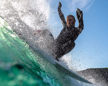 Surfer i svart våtdrakt rir på en bølge med armene hevet mot en blå himmel