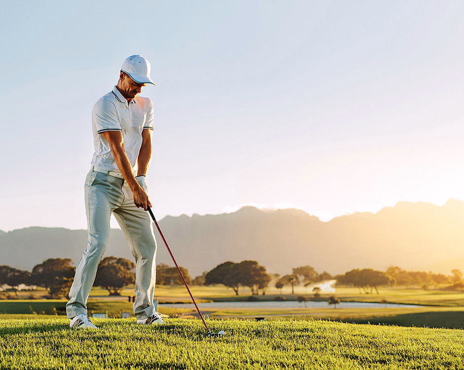 Mann i hvit caps og skjorte klar til å slå en golfball på en bane med fjell bak