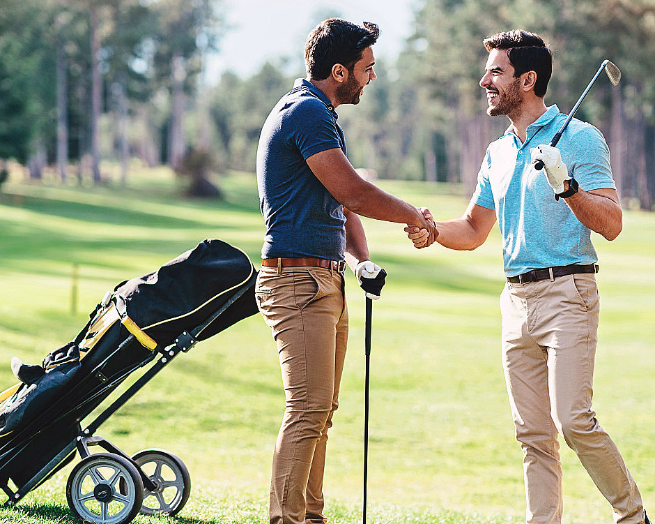 To menn håndhilser på en solfylt golfbane ved siden av en golfbag på tralle