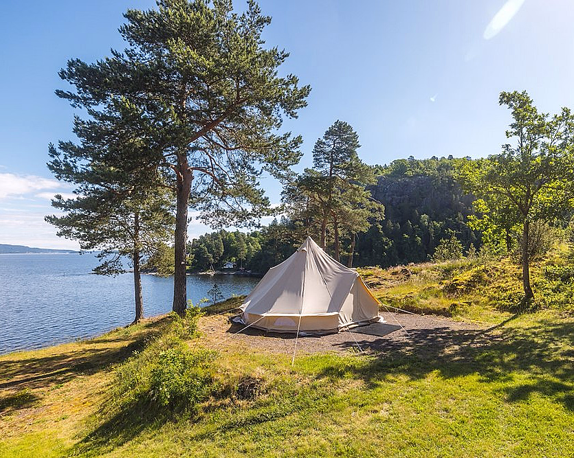 Beige telt på gress ved en innsjø omgitt av furutrær under en blå himmel