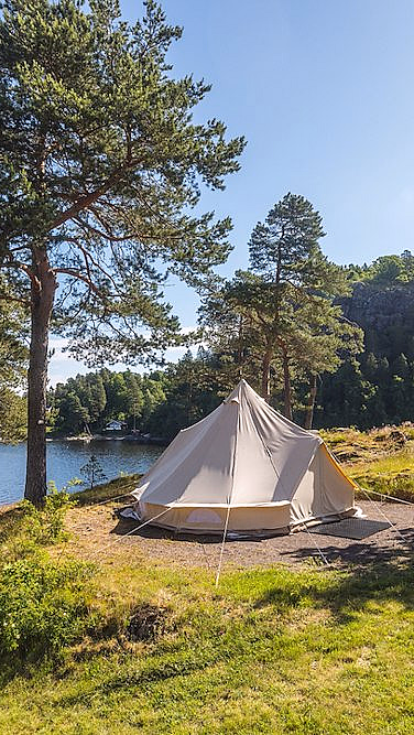 Beige telt på gress ved en innsjø omgitt av furutrær under en blå himmel