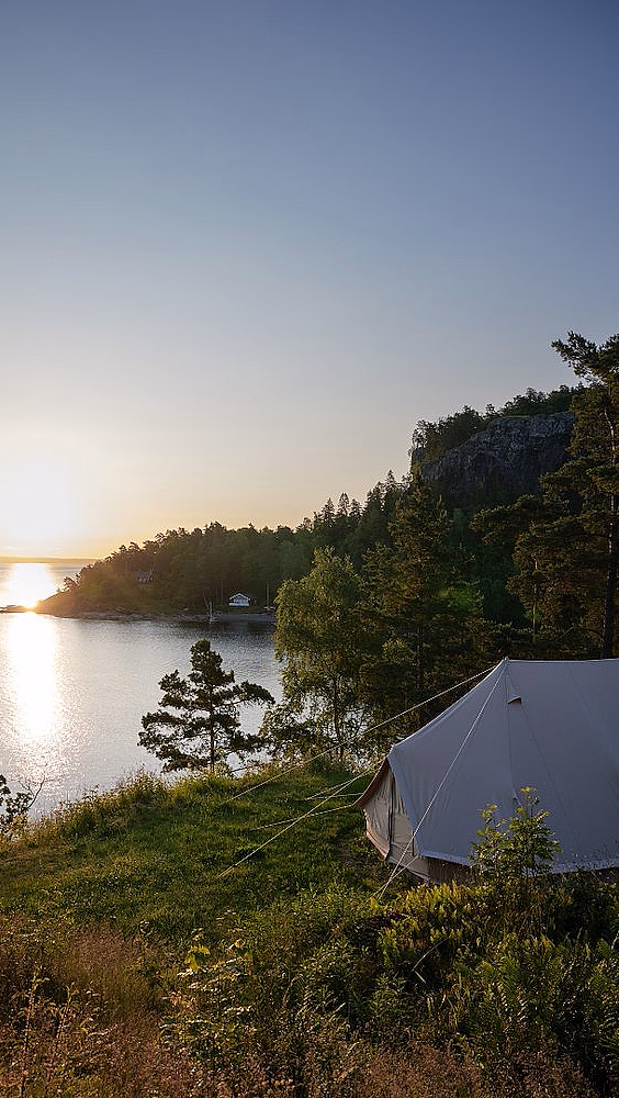 Hvitt telt på gress ved vannet i solnedgang med skog og fjell i bakgrunnen