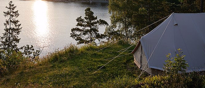 Hvitt telt på gress ved vannet i solnedgang med skog og fjell i bakgrunnen