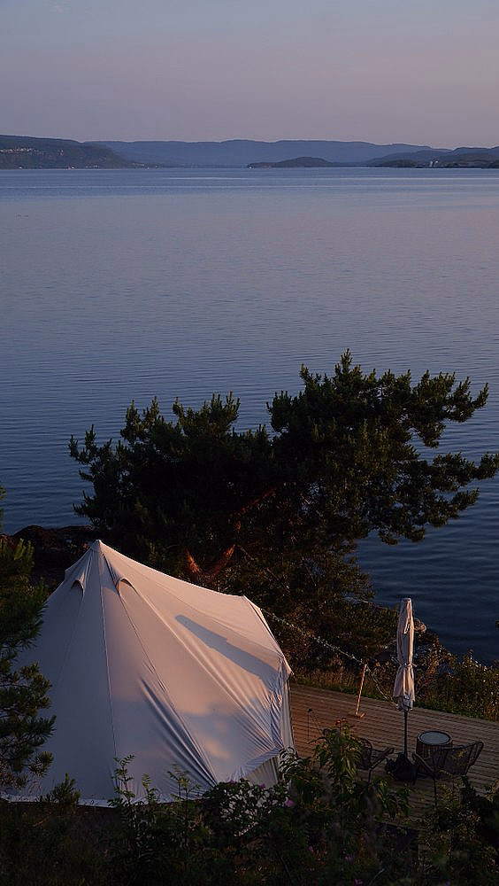 Hvitt telt på treterrasse ved en innsjø med stoler og parasoll i kveldslys