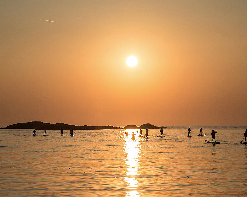Personer på padlebrett i solnedgang med oransje himmel og øyer i bakgrunnen
