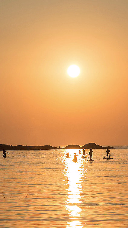 Personer på padlebrett i solnedgang med oransje himmel og øyer i bakgrunnen