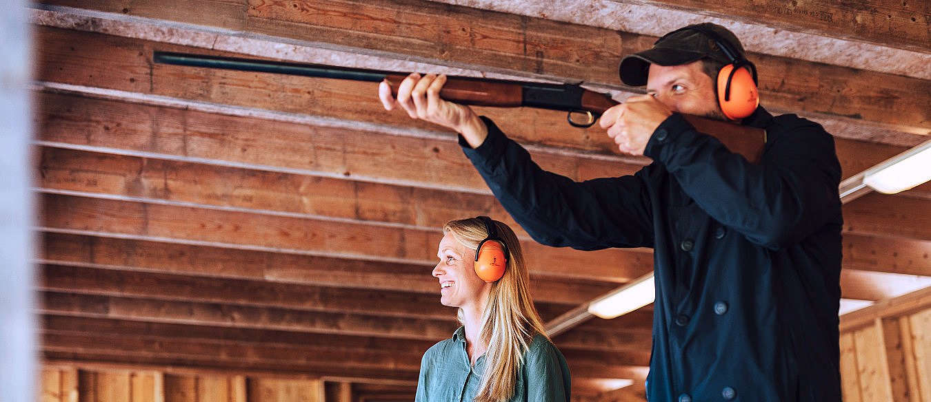 Mann som sikter med rifle og kvinne med oransje hørselvern på en skytebane med trevegger