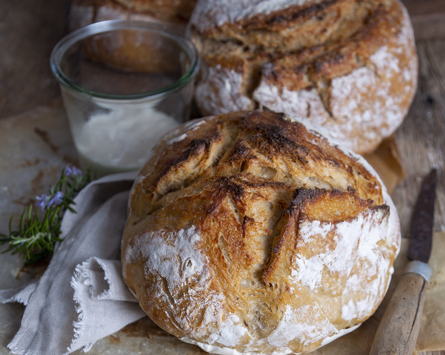 Runde brød med meldryss på bakepapir med en krukke hvit krem, kniv og rosmarin på et trebord
