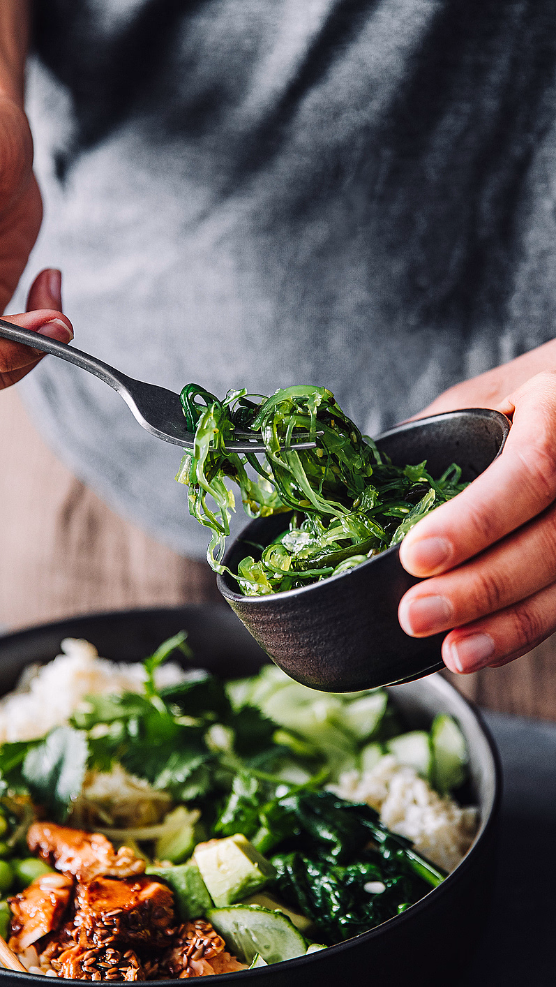 Person som serverer sjøgress i en bolle med ris, avokado, edamame og biter av mat