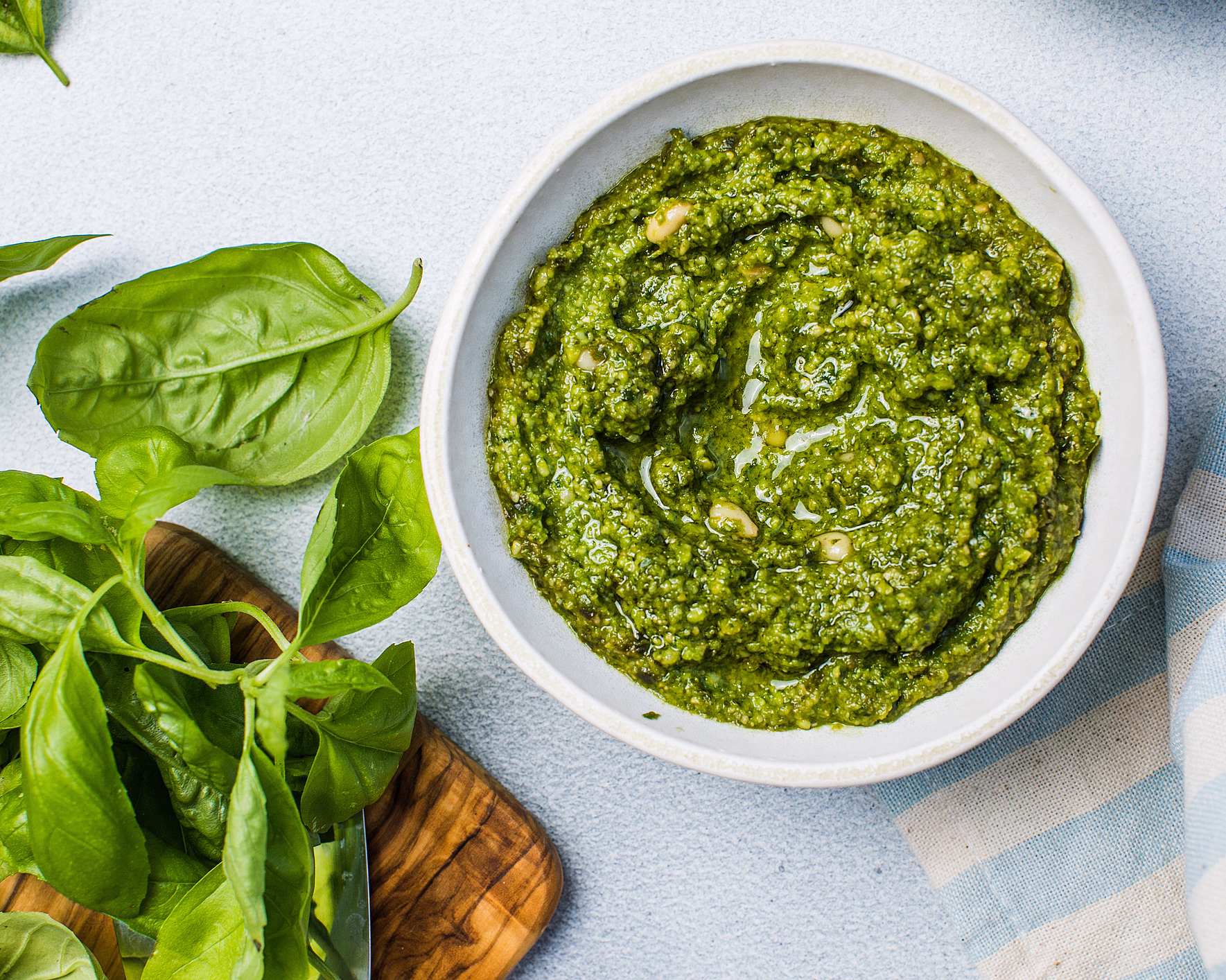 Hvit skål med grønn pesto, basilikumblader på en trefjøl og en blåstripete klut