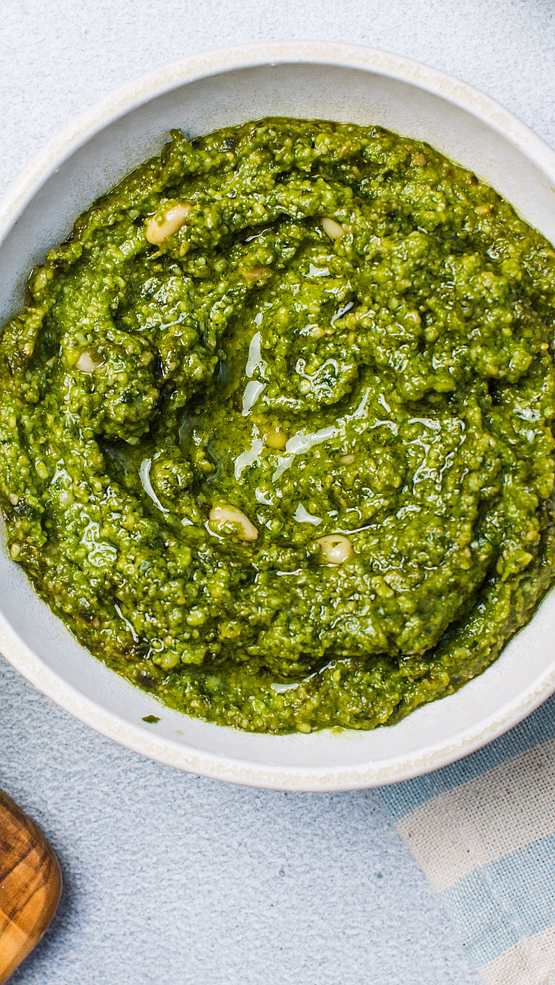 Hvit skål med grønn pesto, basilikumblader på en trefjøl og en blåstripete klut
