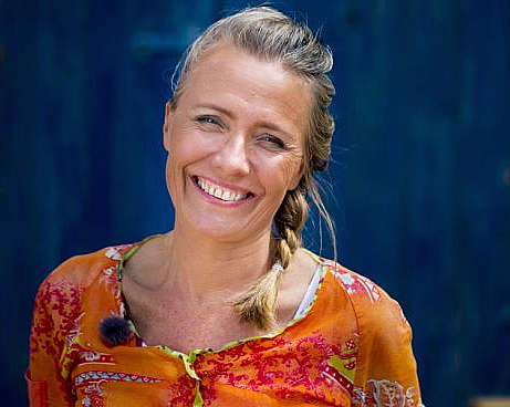 Smilende kvinne med lyst hår i flette og oransje mønstret topp mot blå bakgrunn