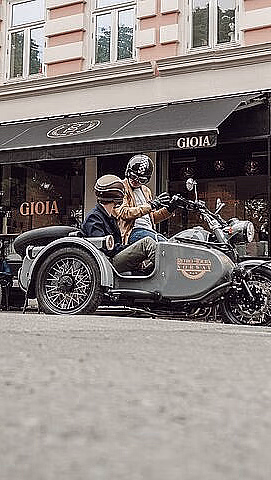 To motorsykler med sidevogner og passasjerer parkert foran en bygning med GIOIA på markisen