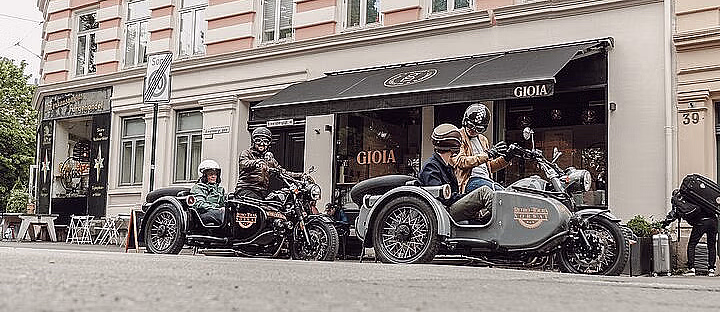 To motorsykler med sidevogner og passasjerer parkert foran en bygning med GIOIA på markisen