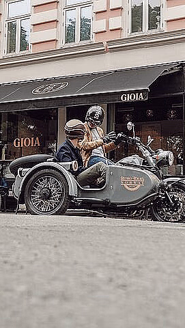 To motorsykler med sidevogner og passasjerer parkert foran en bygning med GIOIA på markisen