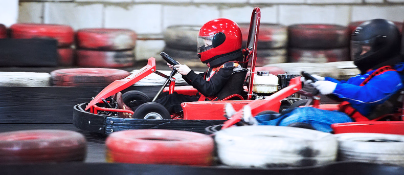 To personer i hver sin gokart på en innendørs bane med dekkstablet langs kanten