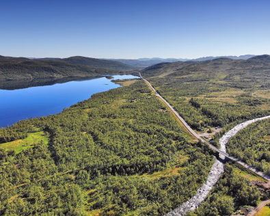 Flyfoto av en vei som strekker seg gjennom et grønt fjellandskap ved siden av en innsjø og en elv