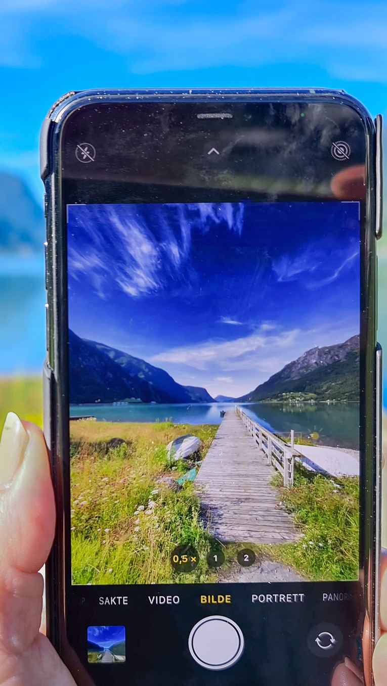 En person holder en mobiltelefon som tar bilde av en brygge ved en fjord omgitt av fjell