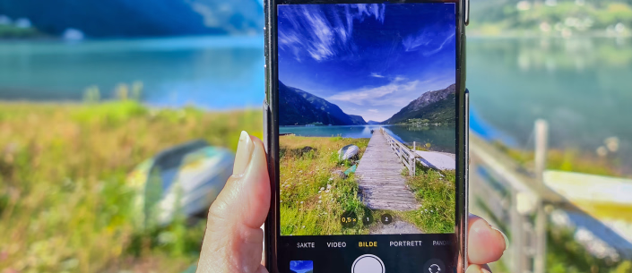 En person holder en mobiltelefon som tar bilde av en brygge ved en fjord omgitt av fjell