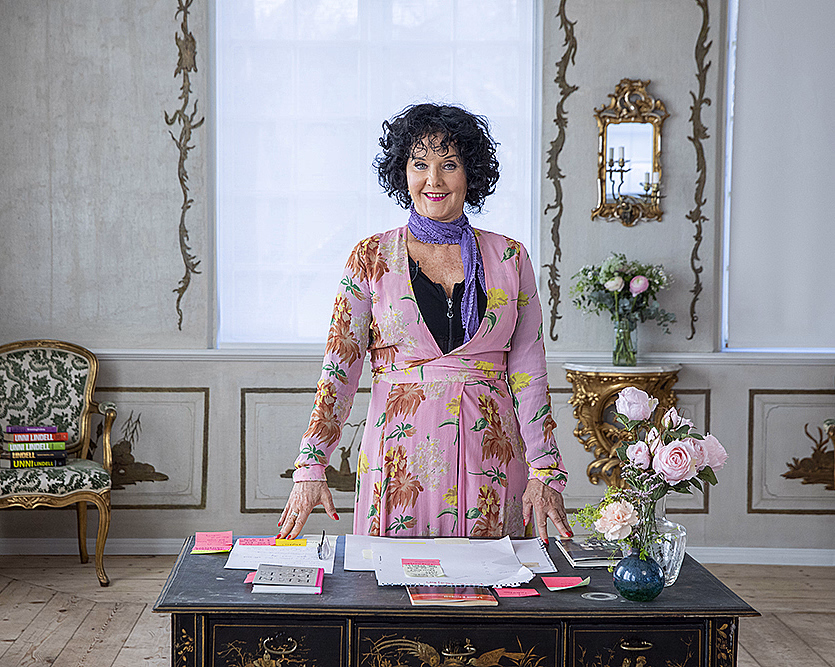 Kvinne i rosa blomsterkjole og lilla skjerf bak et skrivebord i et elegant rom