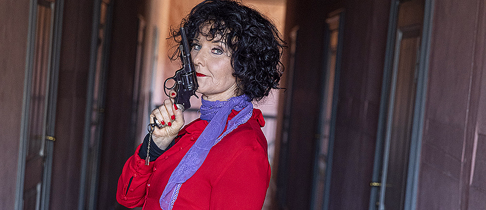 Person med svart krøllete hår i rød kåpe og lilla skjerf holder en revolver i en gang