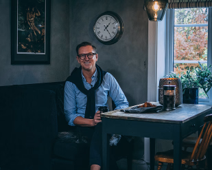 Mann med briller sitter ved et bord med kaffe og en croissant i et rom med en klokke på veggen