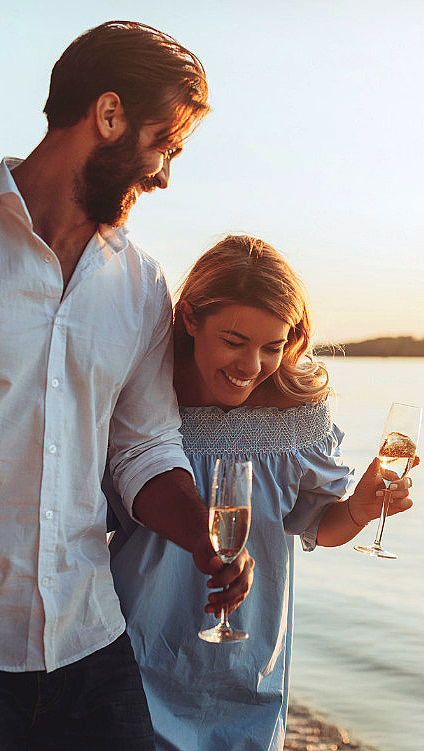 Mann og kvinne som går på en strand ved solnedgang og holder vinglass