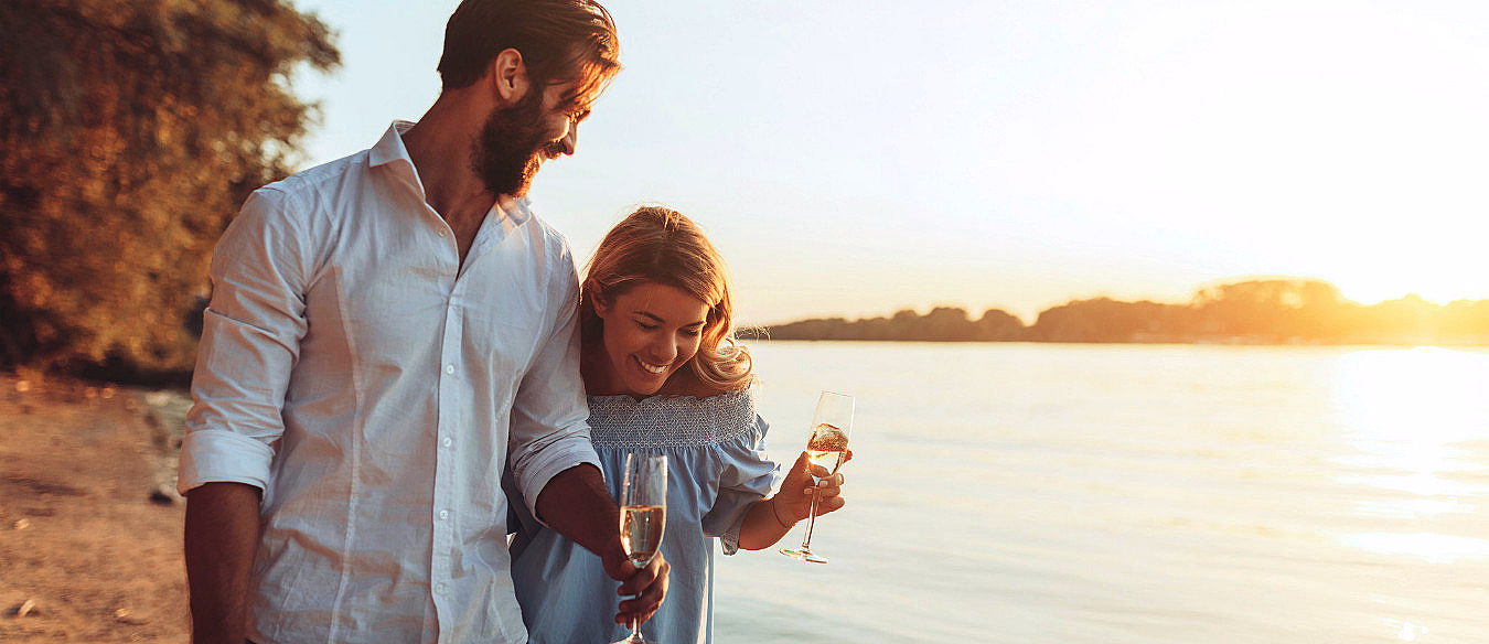 Mann og kvinne som går på en strand ved solnedgang og holder vinglass