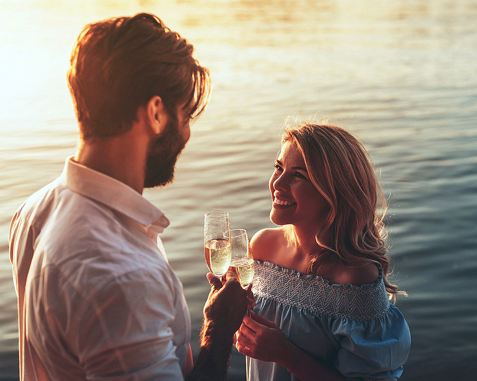 Mann og kvinne ved vannet med champagneglass i solnedgang