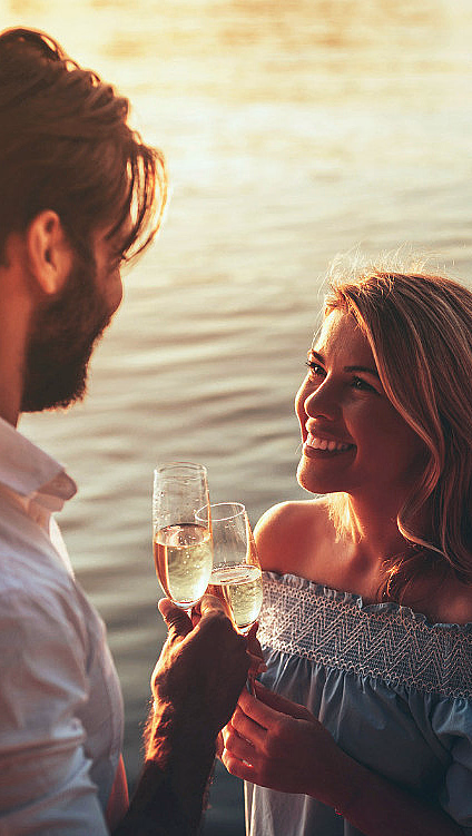 Mann og kvinne ved vannet med champagneglass i solnedgang