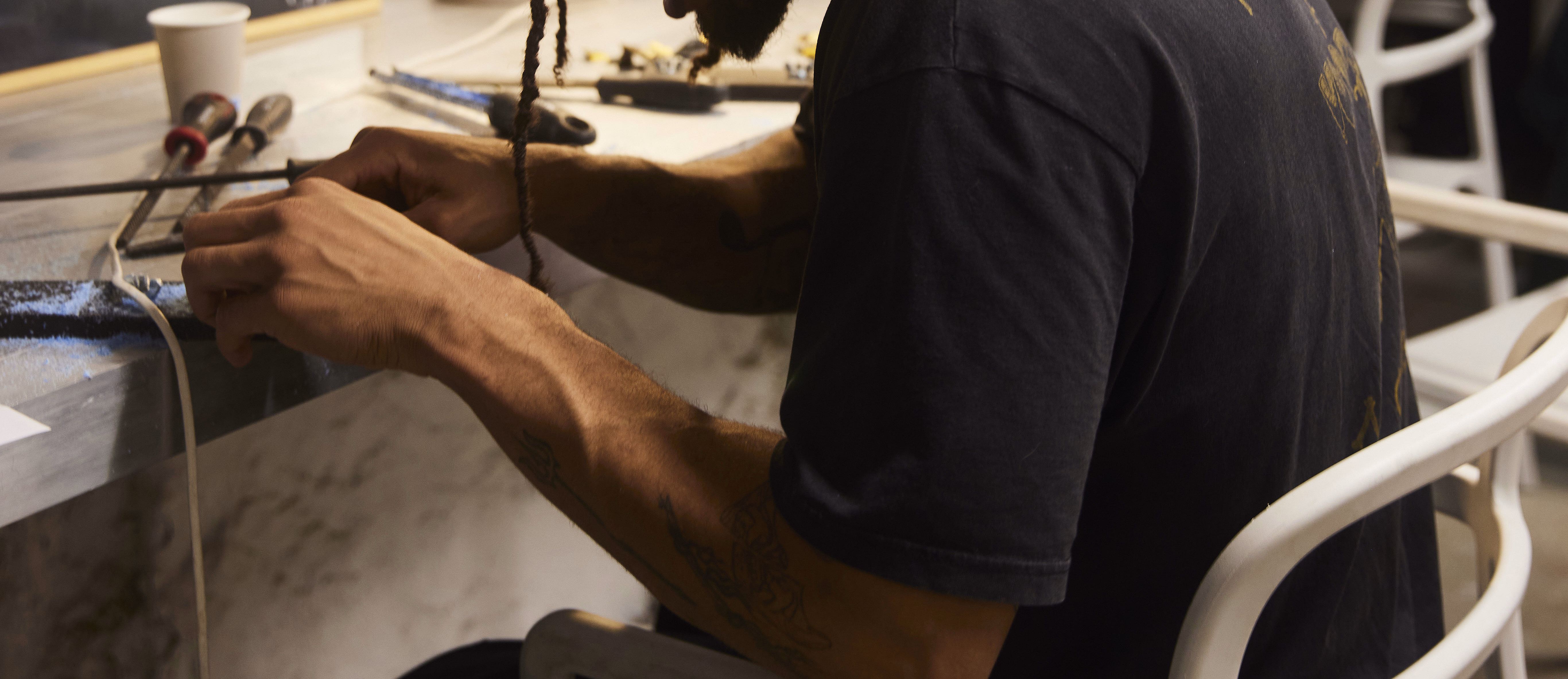 Person med dreadlocks som jobber ved en arbeidsbenk med verktøy og et neonskilt på veggen