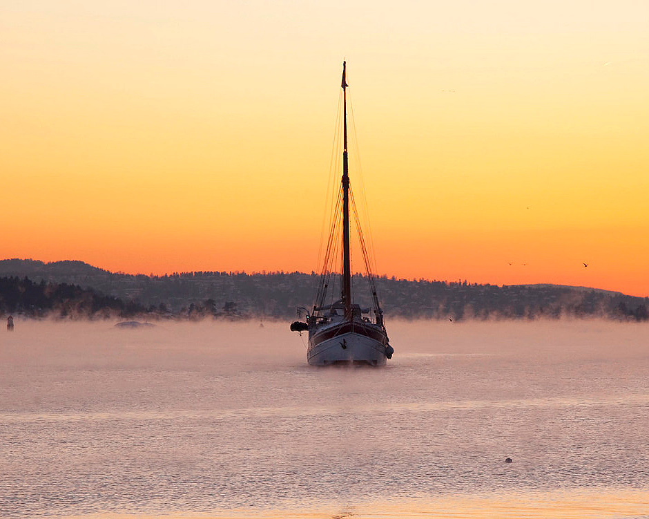 Seilbåt på rolig vann i tåke under en oransje himmel med mørke åser i bakgrunnen