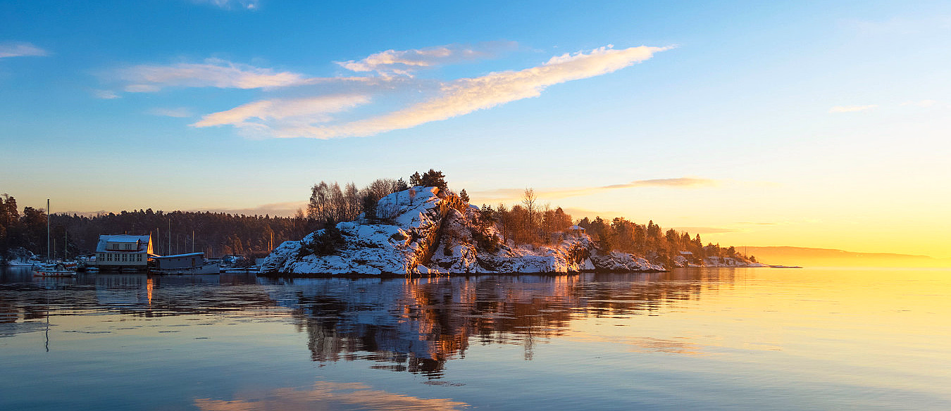 Snødekt øy og et lite hus ved en rolig innsjø i gyllent lys
