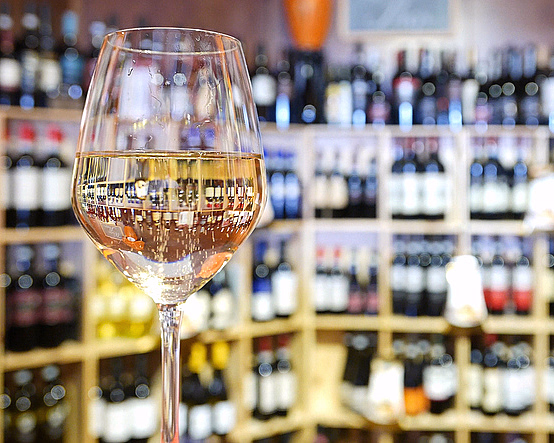 Vinglass med hvitvin foran en trehylle med vinflasker