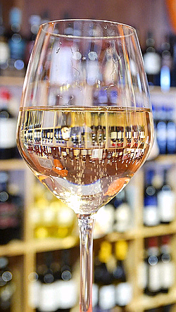 Vinglass med hvitvin foran en trehylle med vinflasker