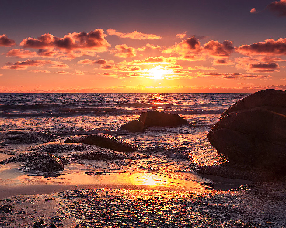 Solnedgang over havet med oransje og lilla himmel, mørke skyer og våte steiner i forgrunnen