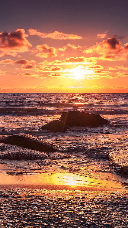 Solnedgang over havet med oransje og lilla himmel, mørke skyer og våte steiner i forgrunnen