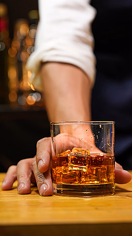 Person i hvit skjorte og mørk vest holder et glass med ravfarget drikke og isbiter