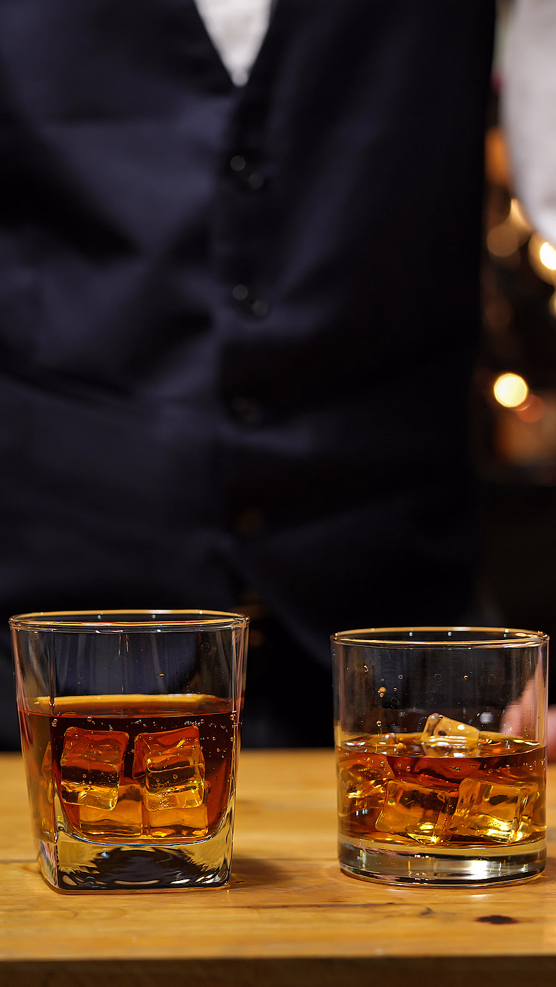 To glass med ravfarget væske og isbiter på en bardisk i tre med en person bak